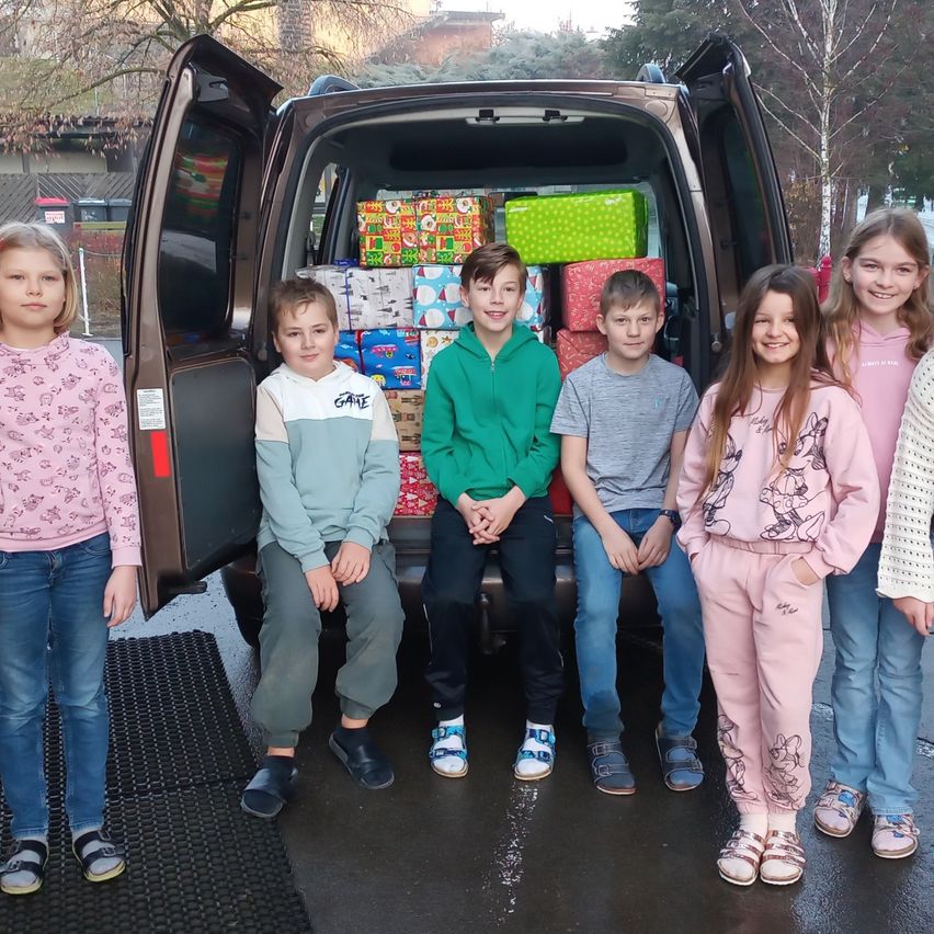 Mehrere Kinder stehen neben einem Minivan mit offenen Hecktüren, gefüllt mit verpackten Geschenken. Sie sind in Winterkleidung gekleidet und posieren wahrscheinlich für ein Foto. Der Hintergrund zeigt Bäume und ein Gebäude.