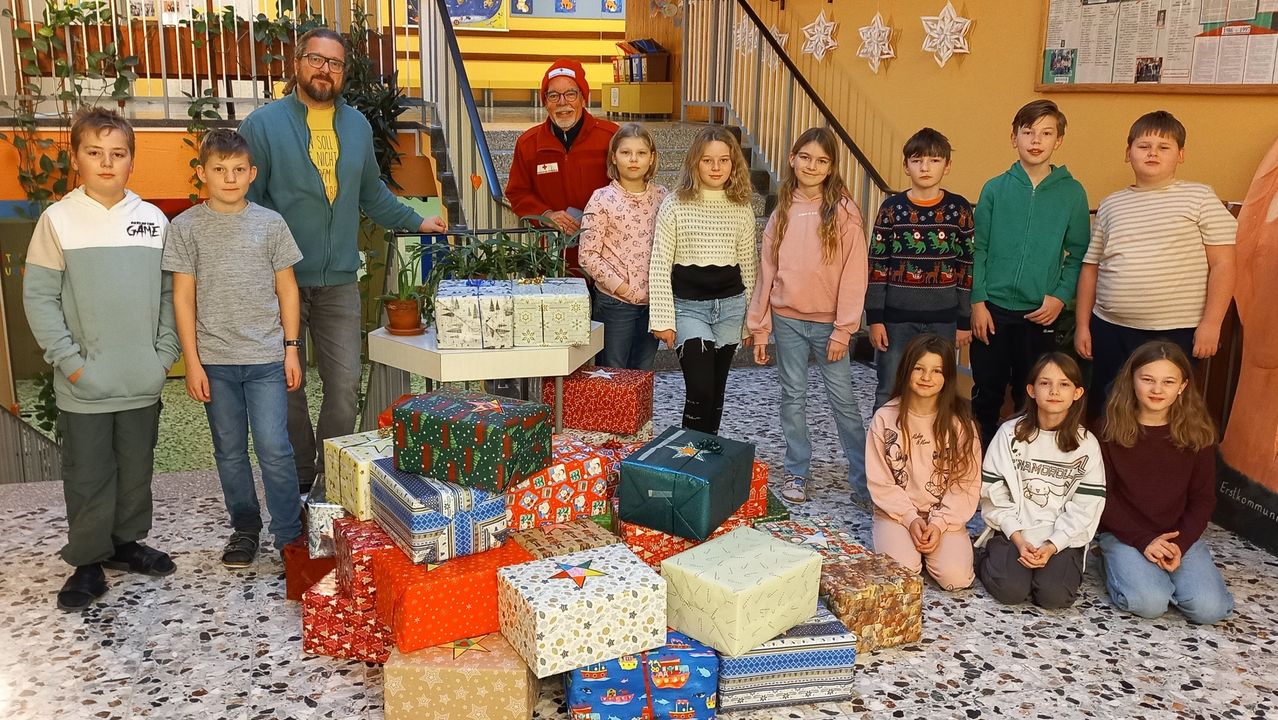 Eine Gruppe von Menschen, einschließlich Kinder, steht vor einer Treppe. Ein Mann in einem roten Mantel ist neben einem Haufen Geschenke. Ein Tisch mit Pflanzen steht dahinter, und Weihnachtsdekorationen hängen an der Wand.