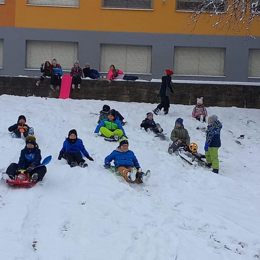 Mehrere Kinder sitzen auf Schlitten in einem verschneiten Bereich vor einem Gebäude mit Fenstern. Einige sitzen im Schnee, während andere von der Mauer des Gebäudes zusehen.