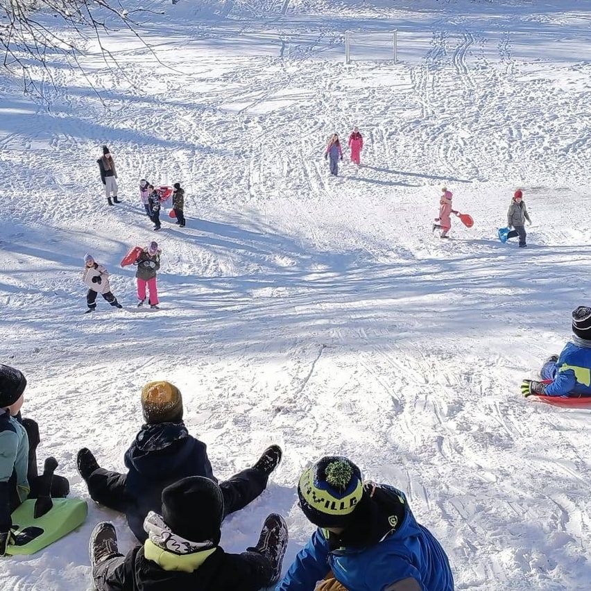 Eine Gruppe von Kindern spielt im Schnee, einige sitzen auf Schlitten, andere laufen oder rennen. Schnee ist auf dem Boden verstreut, und ein Fußballtor ist in der Ferne sichtbar.