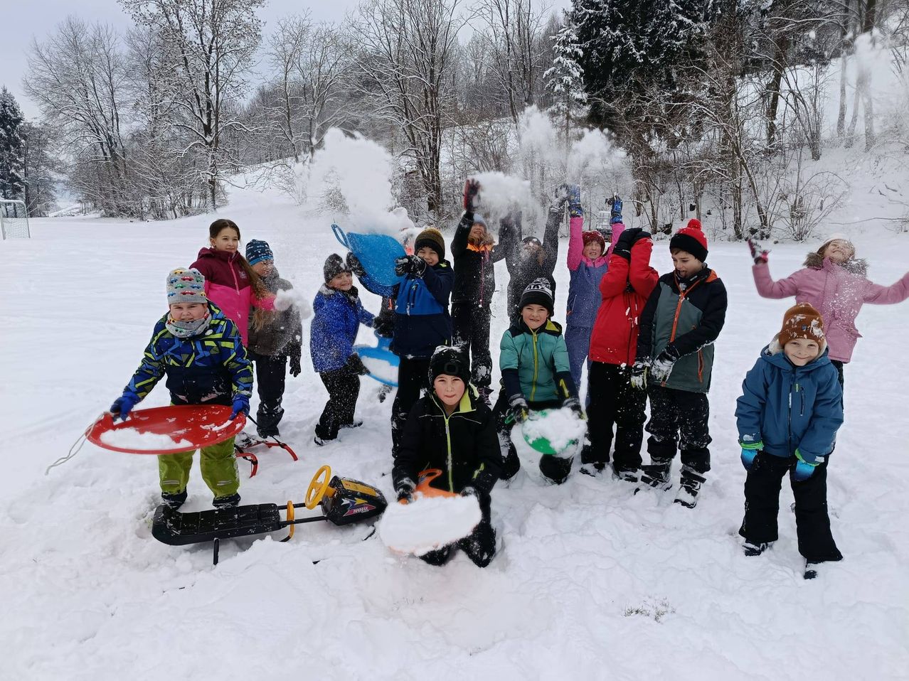 Eine Gruppe von Kindern spielt im Schnee mit Schaufeln und Schlitten, wirft Schnee und einige stehen und andere knien. Sie tragen Winterkleidung und Mützen.