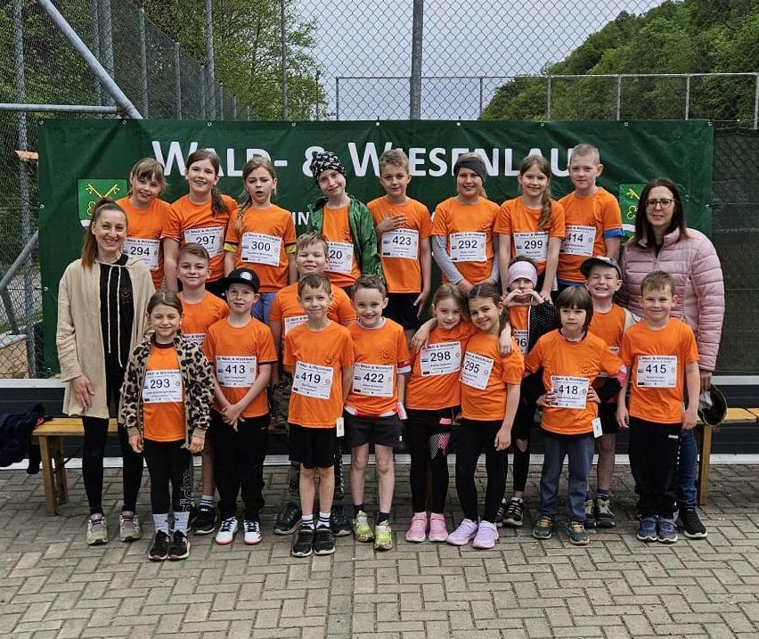 Gruppenfoto von Kindern in orangefarbenen Trikots mit Nummern, die in einer Reihe stehen. Sie posieren für ein Foto vor einem Banner mit grünem Hintergrund. Dahinter steht eine Frau in der Nähe einer Holzbank.
