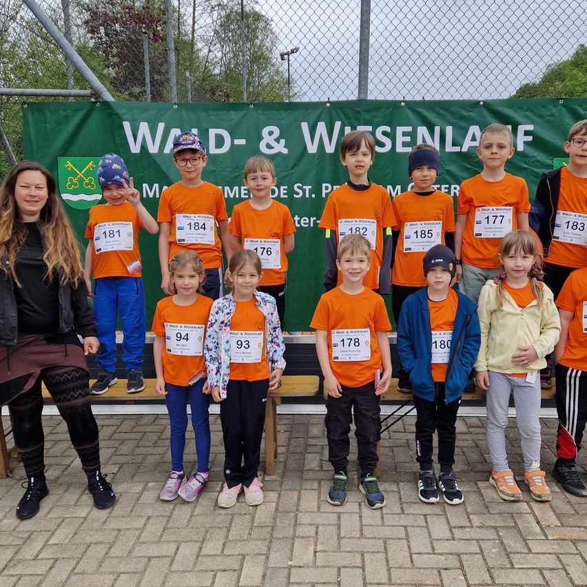 Eine Gruppe von Kindern und Erwachsenen steht zusammen vor einem Banner mit dem Text 'WALD- & WISSENLAF'. Jedes Individuum trägt eine Nummer auf ihren orangefarbenen Shirts.