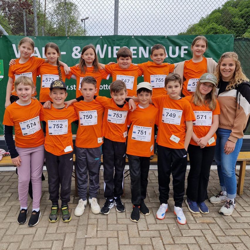 Eine Gruppe von Kindern in orangefarbenen Trikots mit Nummern posiert für ein Foto bei einem Laufevent. Sie lächeln und stehen zusammen, wobei zwei Erwachsene an den Seiten stehen.
