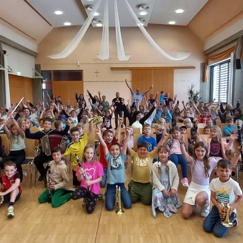 Eine große Gruppe von Kindern mit Musikinstrumenten, sitzend und stehend, feiert in einem Saal. Einige Kinder halten Trophäen. Hinter ihnen steht ein Erwachsener mit erhobenen Händen.