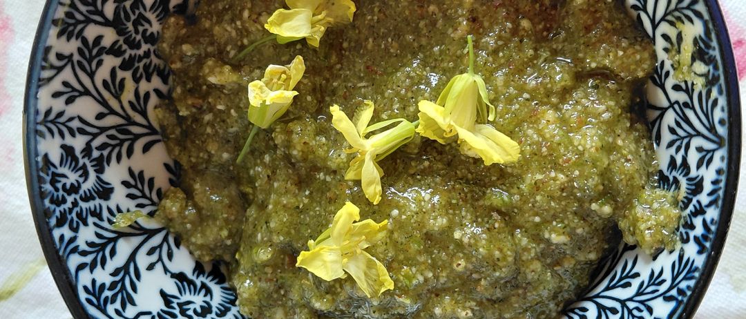 Eine Schüssel mit grüner Paste, verziert mit gelben Blumen, steht auf einer Tischdecke mit Blumenmuster. Die Schüssel hat ein blau-weißes Blumenmuster.