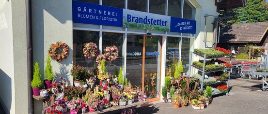 Ein Floristikgeschäft namens Brandstetter mit einer Vielzahl von Blumen und Kränzen vor dem Laden. Das Schild des Geschäfts lautet GARTNEREI BLUMEN & FLORISTIK BRANDSTETTER.