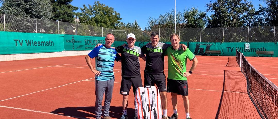 Vier Männer posieren für ein Foto auf einem Tennisplatz, zwei mit Armen umeinander. Eine Tennistasche ruht zwischen zwei von ihnen. Dahinter steht ein Zaun mit grünen Bannern. Bäume und eine Straßenlaterne sind im Hintergrund zu sehen.
