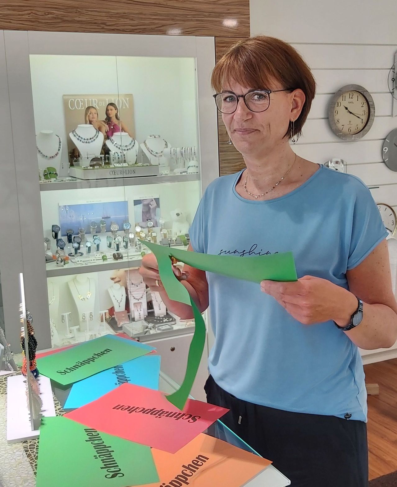 Eine Frau steht vor einem Juwelierladen und hält eine grüne Papierschleife. Sie trägt ein blaues Shirt und eine Brille. Der Laden hat Schmuckdisplays und ein Schild mit der Aufschrift 'COUR DE LION'.