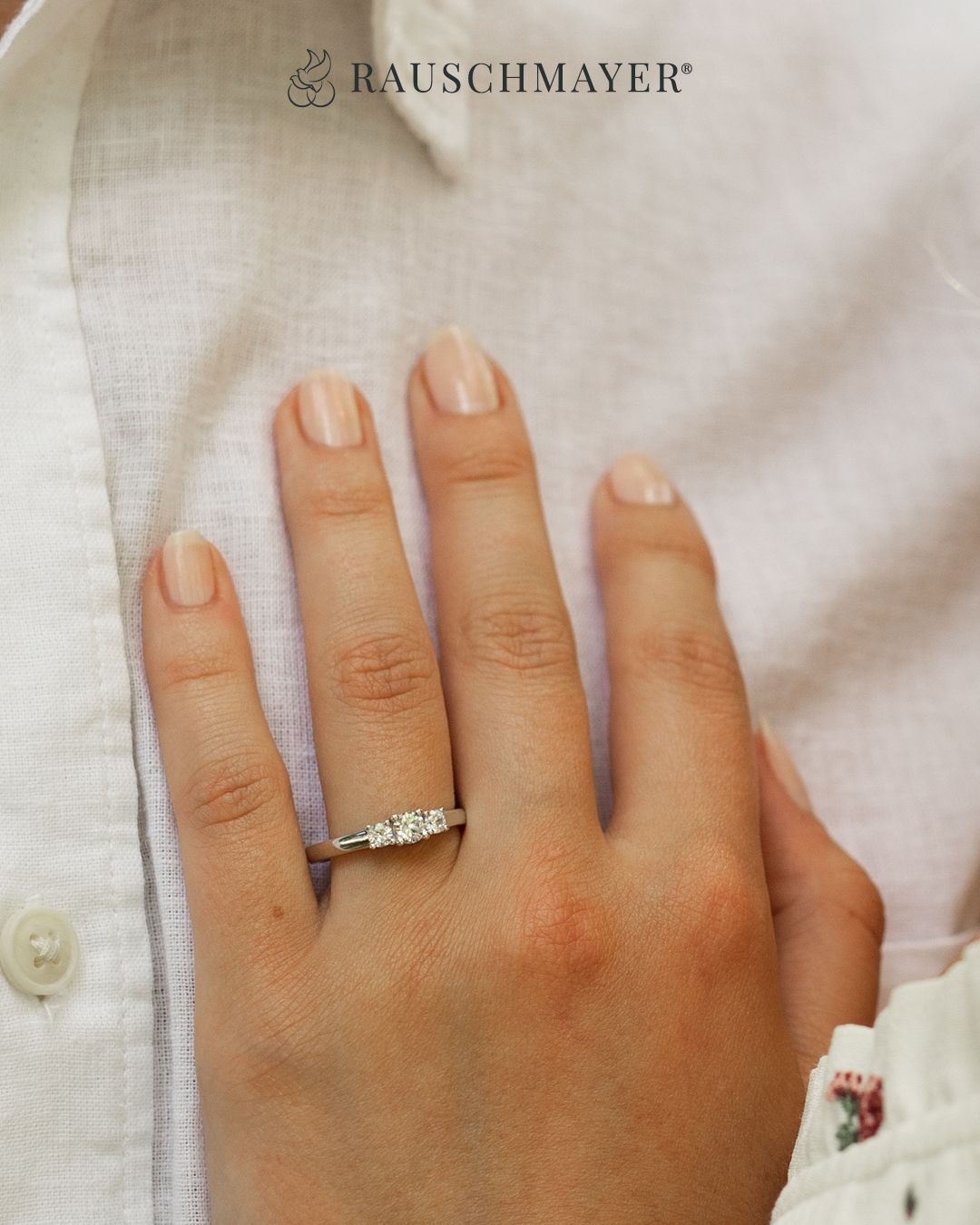 Ein Nahaufnahme einer Hand mit einem drei Stein Diamantring am Ringfinger. Die Hand ist mit einem weißen Hemd geschmückt.