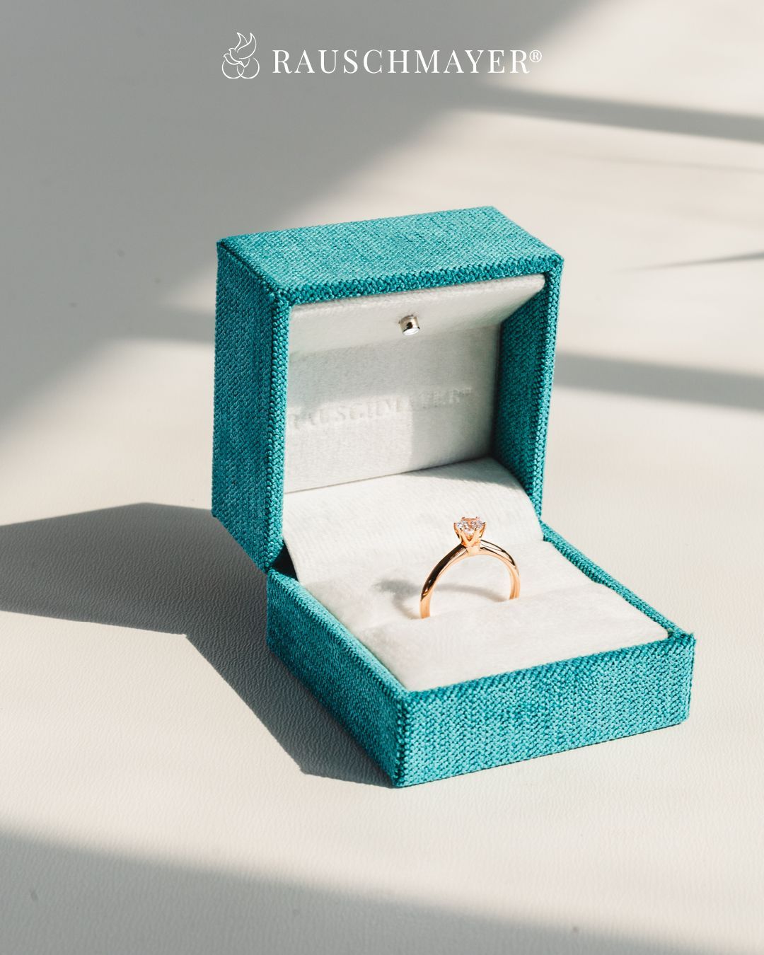 Eine türkisfarbene Samt-Schmuckschatulle enthält einen Roségold-Verlobungsring mit einem zentralen Diamanten, der auf einem weißen Samtkissen ruht.