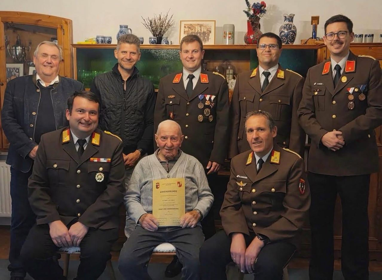 Eine Gruppe von Militärangehörigen posiert für ein Foto, wobei ein Mann einen Ausweis in der Hand hält. Sie alle lächeln und scheinen sich in einem Raum zu befinden.