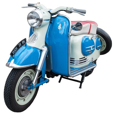 Ein altmodischer Roller mit blauer und weißer Lackierung, Scheinwerfer und einem Nummernschild, das 'DUCH SR 1190' liest. Er hat einen kleinen Reservereifen und einen Metallständer.