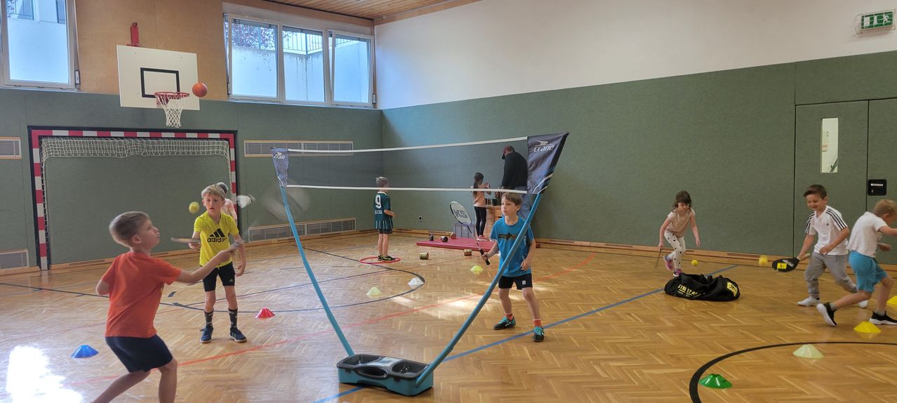 Eine Gruppe von Kindern spielt Badminton in einer Turnhalle. Ein Junge in einem blauen Shirt schlägt den Federball über das Netz mit einem Schläger. Andere Kinder stehen mit Schlägern herum. Die Halle hat einen Holzboden und grüne Wände.