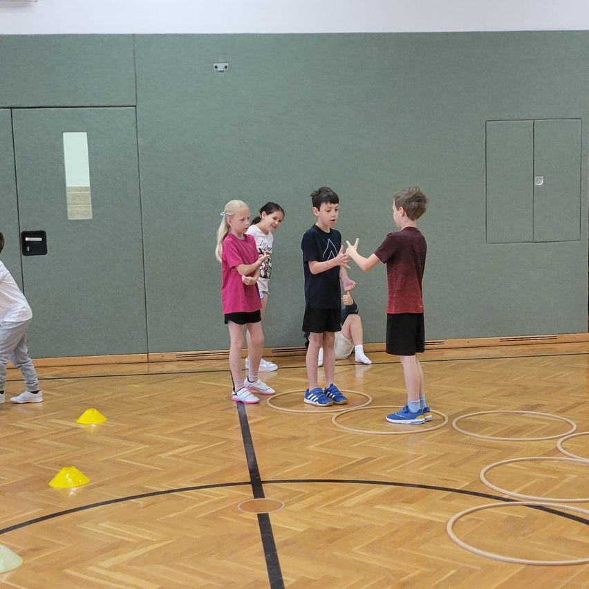Eine Gruppe von Kindern in einer Turnhalle steht im Kreis. Einige interagieren, während andere zusehen. Gelbe Kegel sind um sie herum platziert.