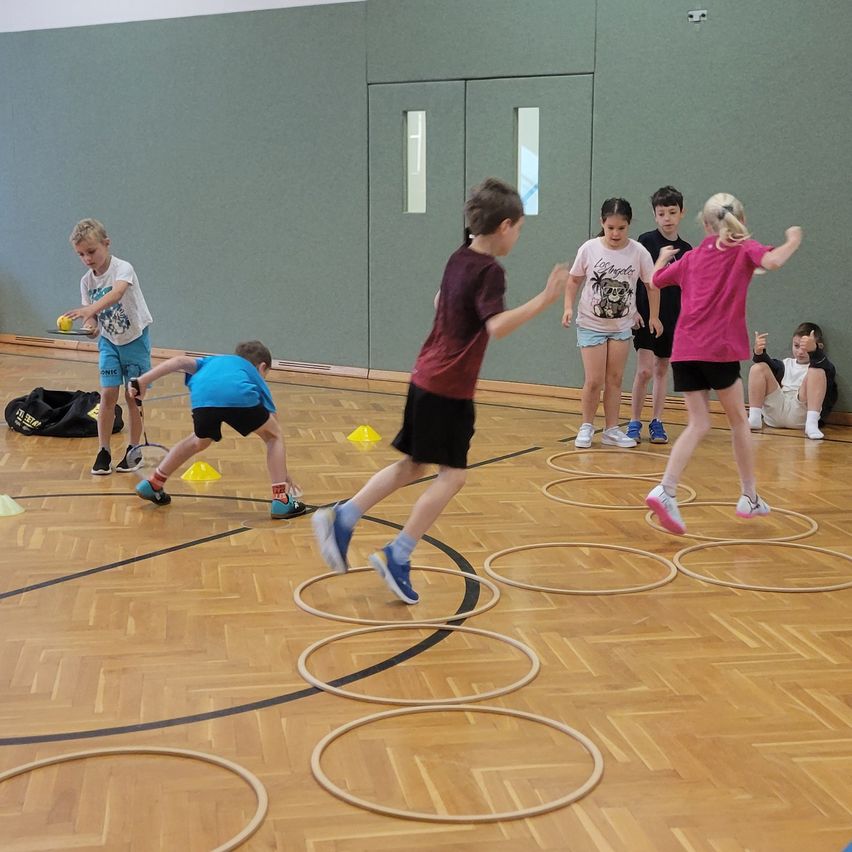 Mehrere Kinder spielen ein Geschicklichkeitsspiel auf einem Hallenplatz und navigieren durch Reifen, während einige gelbe Bälle halten.