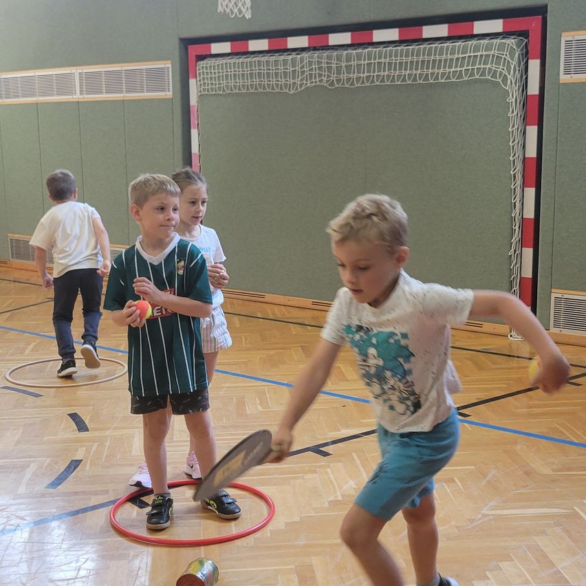 Eine Gruppe von Kindern spielt mit Schlägern und Bällen in einer Turnhalle. Ein Kind ist dabei, den Ball zu schlagen, während zwei andere zuschauen. Im Hintergrund steht ein Fußballtor.