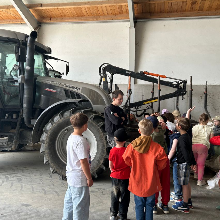 Eine Gruppe von Kindern, begleitet von einem Mann, steht vor einem Traktor in einer Scheune. Der Mann erklärt ihnen etwas.