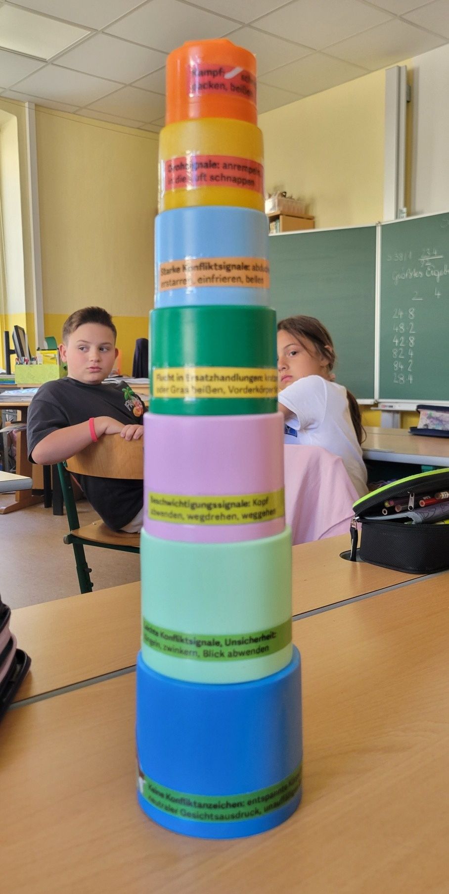 Zwei Kinder sitzen in einem Klassenzimmer mit einem Stapel farbiger Zylinder im Vordergrund. Der Junge trägt ein schwarzes T-Shirt, und das Mädchen hat ein pinkes Shirt. Dahinter befindet sich eine Tafel mit Zahlen.