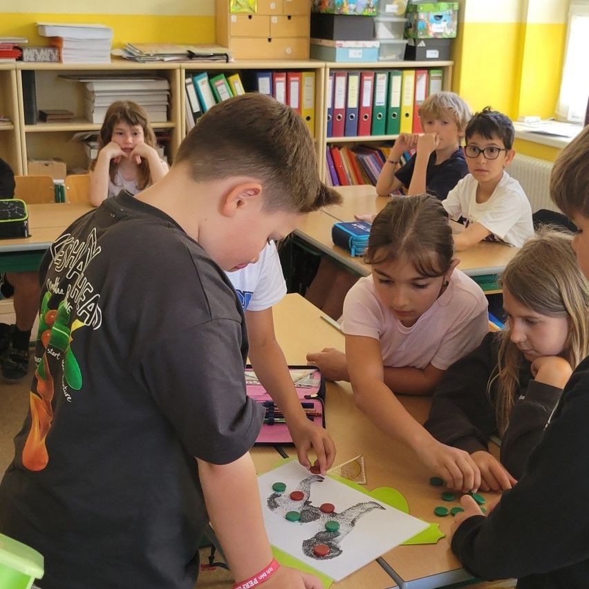 Ein Klassenzimmer mit Kindern, die an Tischen sitzen und an einem Projekt arbeiten. Ein Junge in einem schwarzen Shirt zeichnet auf einem Blatt Papier, während andere verschiedene Aktivitäten verfolgen.