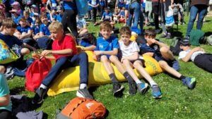 Mehrere Kinder sitzen auf einem gelben Schlauchboot auf dem Rasen, einige sitzen auf dem Boden. Einige der Kinder tragen Rucksäcke. Hinter ihnen sitzen noch andere Menschen auf dem Rasen.