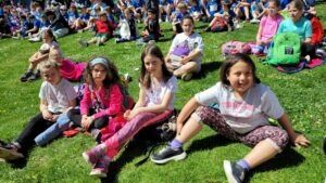 Eine Gruppe von Kindern sitzt auf einem Grasfeld. Sie tragen verschiedene Kleidungsstücke und Schuhe. Einige von ihnen tragen Rucksäcke und Handtaschen. Sie sitzen und lächeln, während sie etwas vor sich anschauen. Hinter ihnen sitzen und stehen viele andere Menschen.