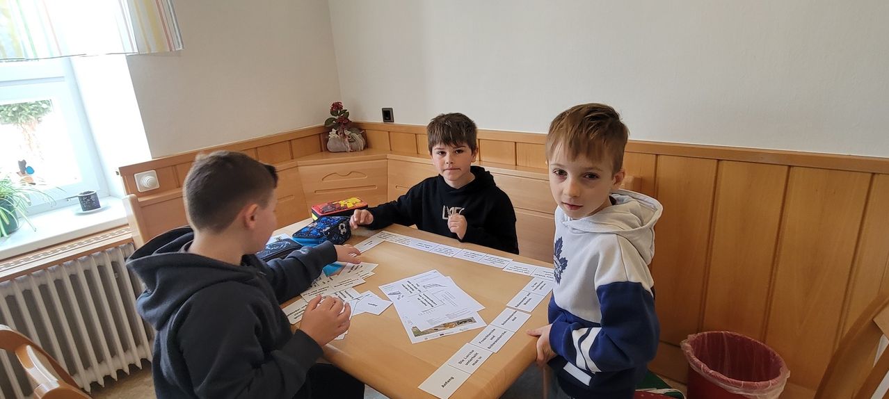 Drei junge Jungen sitzen um einen Tisch mit Karten und Papieren und beschäftigen sich mit einer Aktivität. Der Junge in der Mitte trägt eine schwarze Kapuzenjacke.