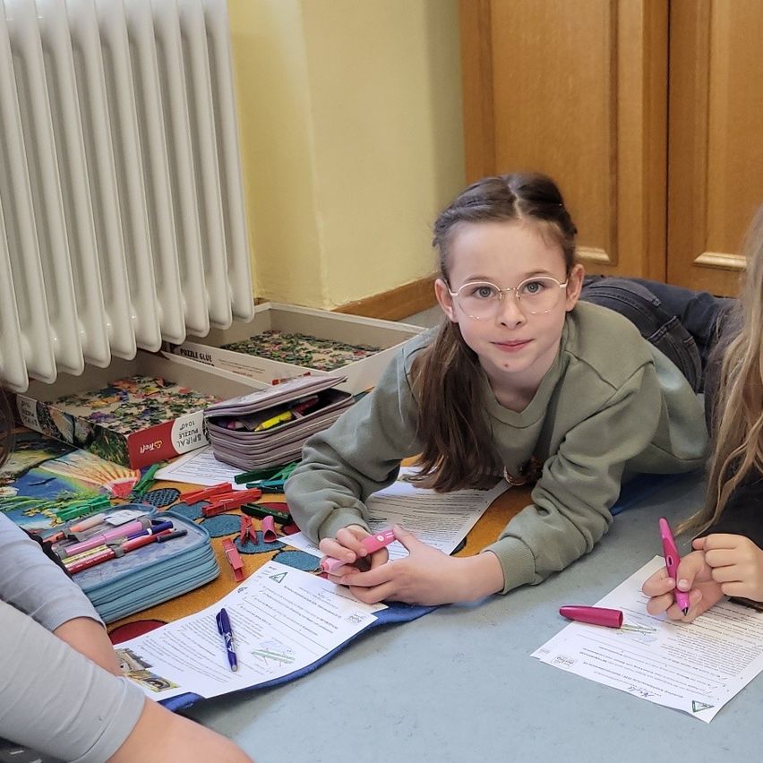 Zwei junge Mädchen liegen auf einem Schreibtisch und beschäftigen sich mit farbigen Markern und Papieren. Ein Puzzlekasten, ein Federmäppchen und eine Heizung sind sichtbar.