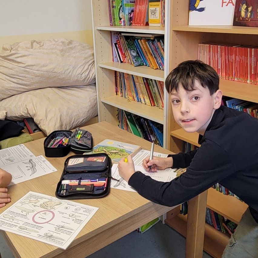 Ein junger Junge sitzt an einem Schreibtisch in einem Klassenzimmer und schreibt mit einem Stift auf Papier. Verschiedene Kunstmaterialien liegen auf dem Tisch. Bücher und andere Gegenstände befinden sich in den Regalen hinter ihm.