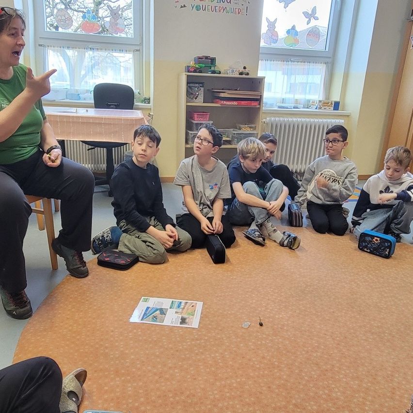 Eine Frau in einem grünen Shirt unterrichtet eine Gruppe von Jungen, die im Klassenzimmer auf dem Boden sitzen. Sie gestikuliert mit der Hand. Ein Heizkörper befindet sich an der Wand hinter ihnen.