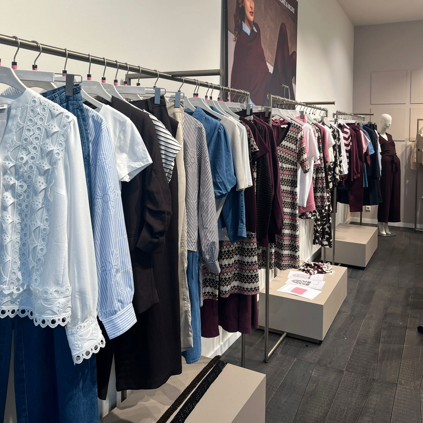 Ein Bekleidungsgeschäft zeigt eine Vielzahl von Kleidungsstücken auf Kleiderständern, darunter Blusen, Kleider und Mäntel. Im Hintergrund befinden sich Mannequins und Poster.