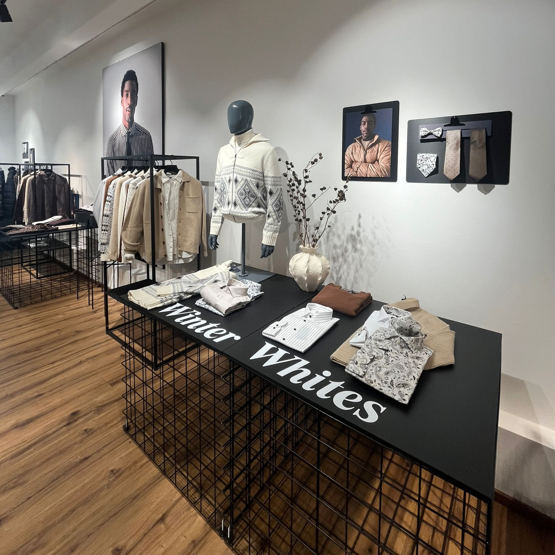 Eine Kleidungsausstellung zeigt einen Mannequin in einem weißen Pullover, einen schwarzen Tisch mit den Worten 'Winter Whites' und verschiedene Kleidungsstücke. Ein Vasen mit einer Pflanze und gerahmte Bilder schmücken die Wand.