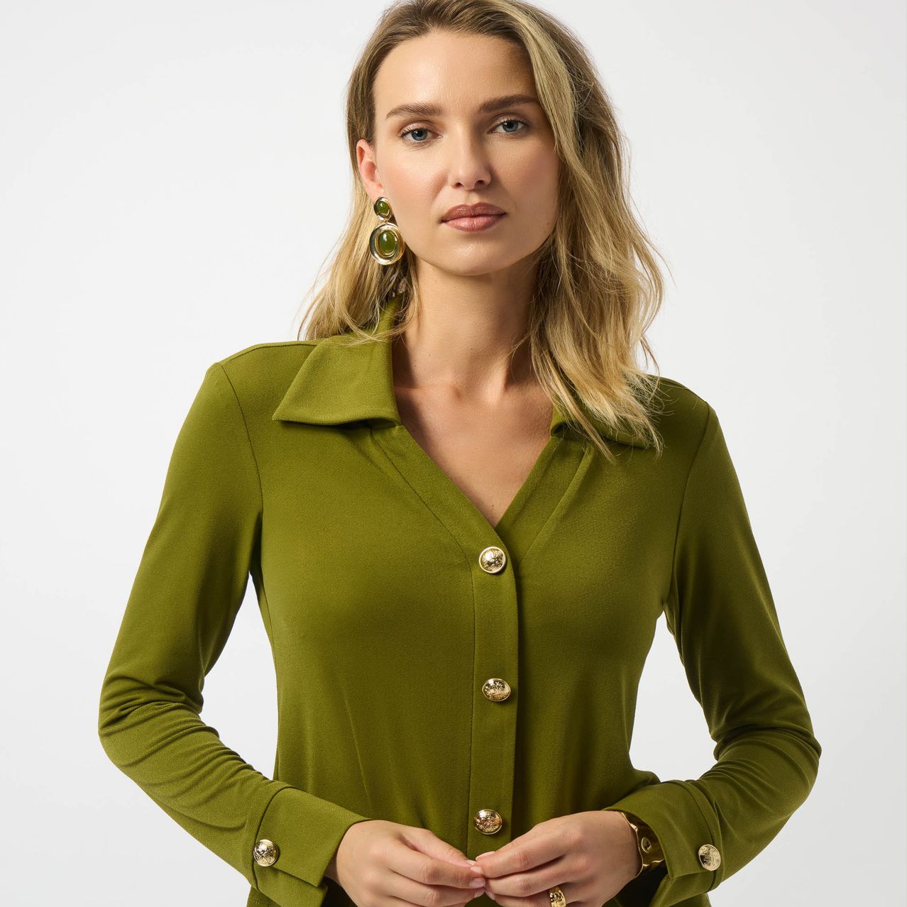 Eine blonde Frau posiert in einem grünen Langarmhemd mit goldenen Knöpfen vor weißem Hintergrund.