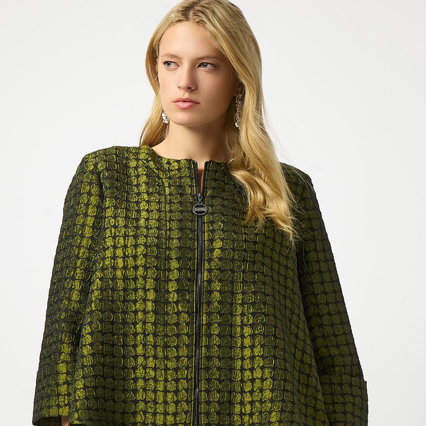 Eine Frau mit blonden Haaren trägt eine grün gemusterte Jacke mit schwarzem Reißverschluss. Sie posiert für ein Foto.