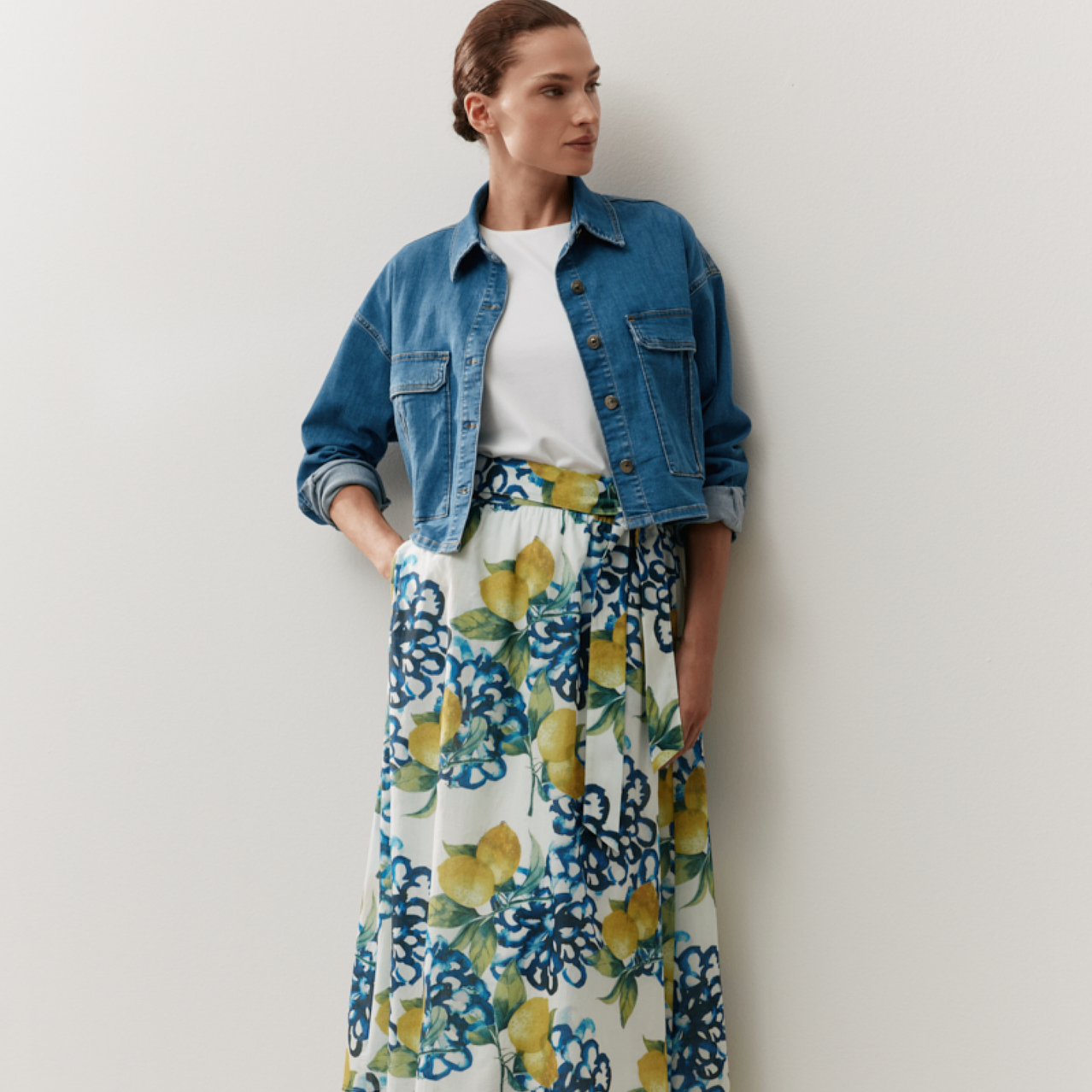 Eine Frau in einem weißen Oberteil, einer Jeansjacke und einem Lemon-bedruckten Rock steht an einer weißen Wand.