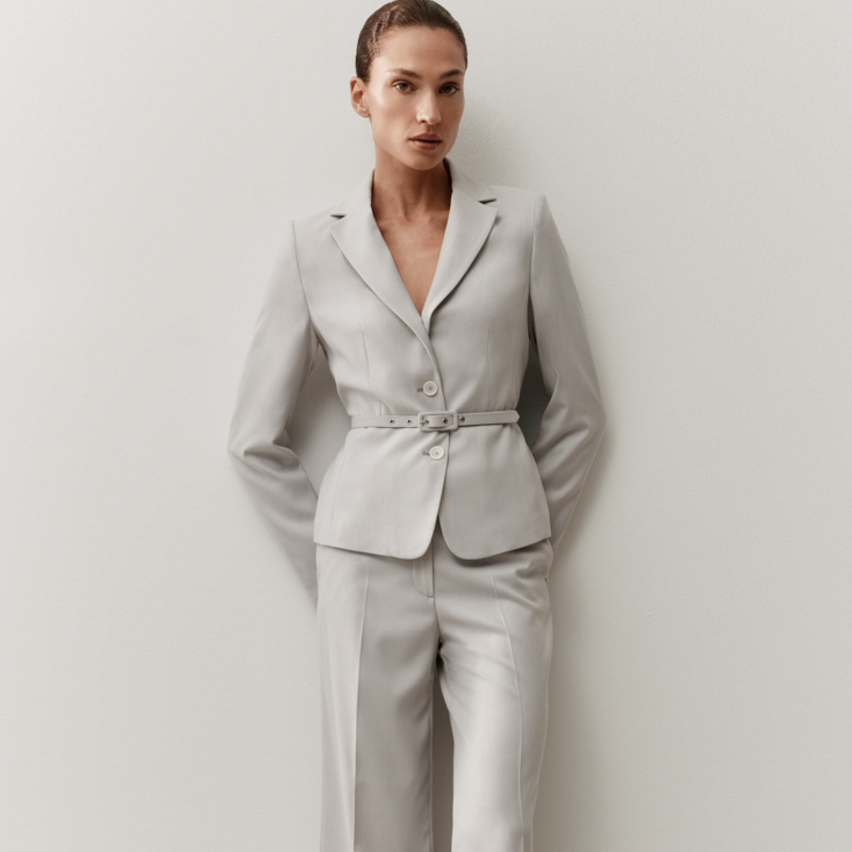 Eine Frau in einem hellgrauen Blazer und dazu passenden Hosen steht an einer weißen Wand.