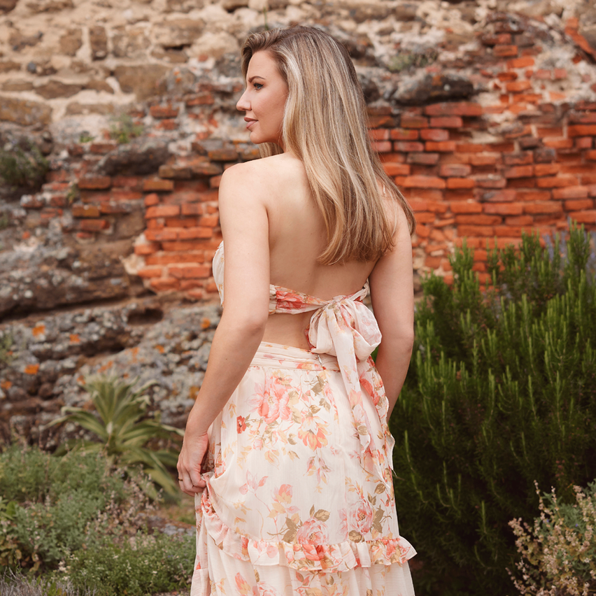 A woman with blonde hair, standing with her back to the camera, wearing a floral dress. Behind her is a brick wall.