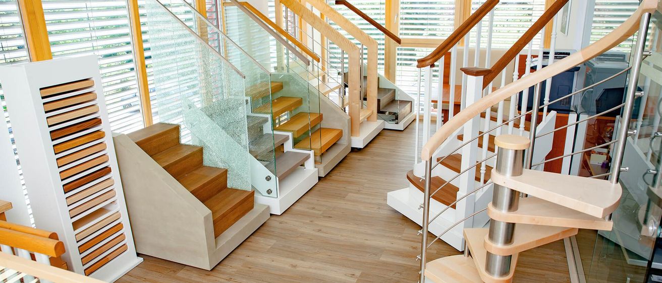 Ein modernes Interieur mit drei verschiedenen Treppen: eine mit Glasstufen und Metallgeländern, eine andere mit Holzstufen und Geländern und eine Wendeltreppe. Alle Treppen stehen an einem Holzboden.