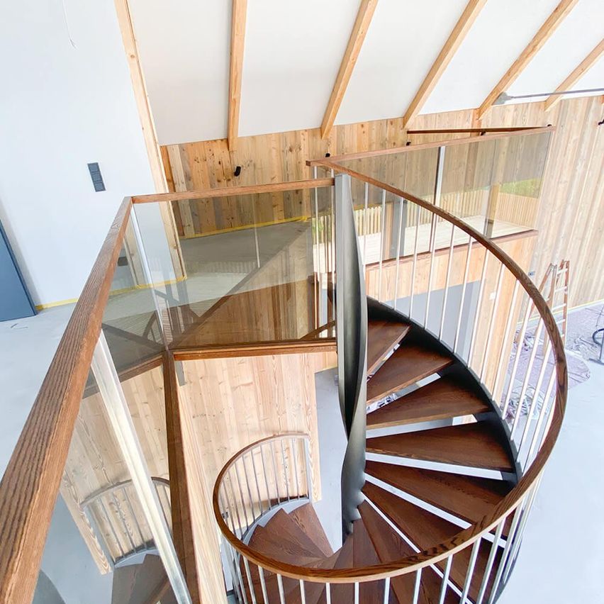 Ein Blick von oben auf eine hölzerne Wendeltreppe mit Glasgeländern und einer hölzernen Decke. Das Haus hat eine Holzwand.