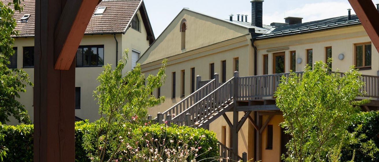 Ein Gartenblick von einer Hausveranda, der ein großes Gebäude mit einer Holztreppe zeigt, umgeben von Grün und Pflanzen.