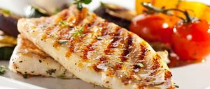 Ein Teller mit zwei gegrillten Fischfilets, garniert mit Kräutern und begleitet von Gemüse wie Auberginen und Tomaten.