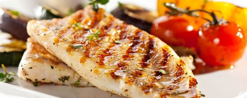 Ein Teller mit zwei gegrillten Fischfilets, garniert mit Kräutern und begleitet von Gemüse wie Auberginen und Tomaten.