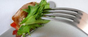 Eine Gabel hält ein Stück gegrilltes Hähnchen mit grünem Salat und Tomaten auf einer weißen Oberfläche.