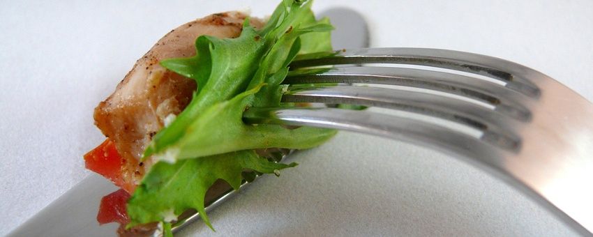 Eine Gabel hält ein Stück gegrilltes Hähnchen mit grünem Salat und Tomaten auf einer weißen Oberfläche.