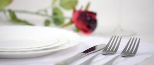 Eine ordentlich gedeckte Tischlandschaft mit weißen Tellern, einem Messer und zwei Gabeln, begleitet von einer roten Rose im Hintergrund.