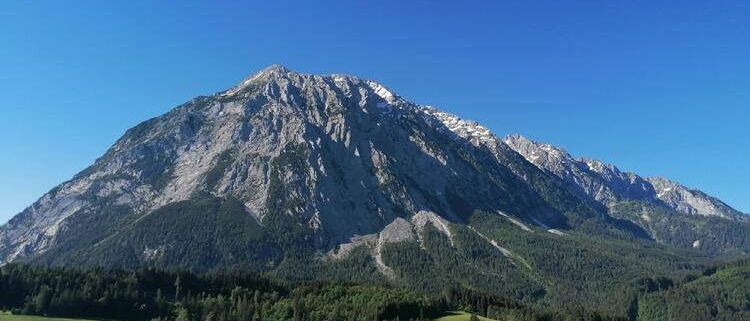 Ein Gebirgszug mit Schnee auf den Gipfeln. Die Berghänge sind mit Bäumen bedeckt. Der Himmel ist blau.