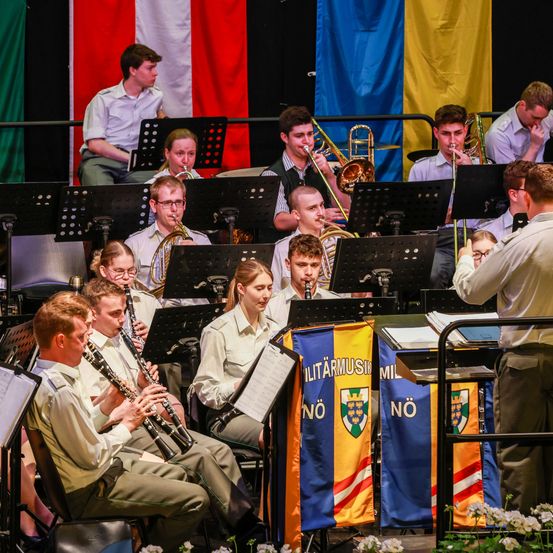 Ein Militärorchester probet auf der Bühne, mit Musikern, die Trompeten, Posaunen und Klarinetten spielen. Der Dirigent steht rechts, und das Orchester ist mit Flaggen geschmückt.