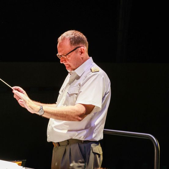 Ein älterer Mann in einem weißen Hemd und mit Brille hält einen Dirigentenstab in der Hand und steht auf einer Bühne. Er scheint eine musikalische Aufführung zu leiten.