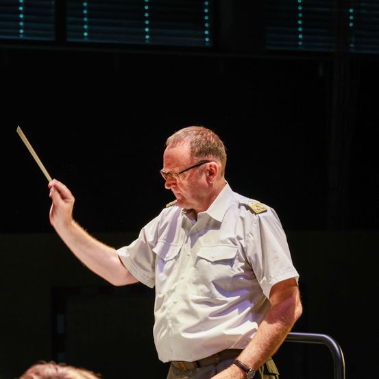 Ein älterer Mann in einem weißen Uniformkleid dirigiert ein Orchester mit einem Taktstock. Die Szene ist schwach beleuchtet.