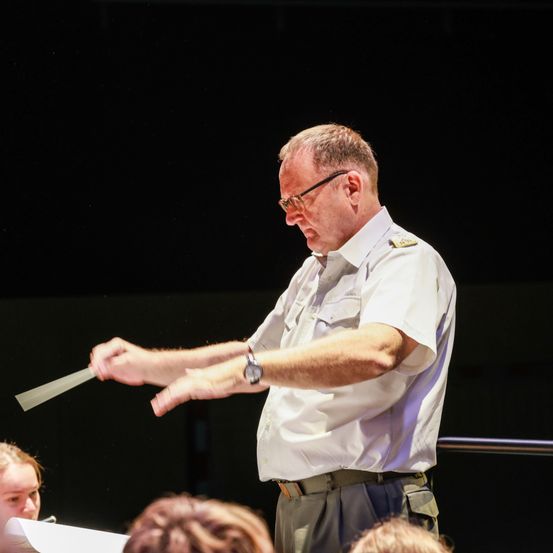 Ein erwachsener Mann in weißer Uniform mit Taktstock dirigiert ein Orchester auf der Bühne. Das Publikum ist nicht sichtbar.